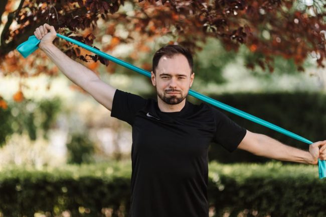 Adult man in black shirt exercises with a resistance band outside.
