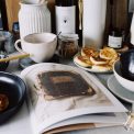 One portion of homemade cheese pancakes in frying pan and another on plate locating near opened stylish magazine among kitchen utensils and appliances in bright kitchen at home