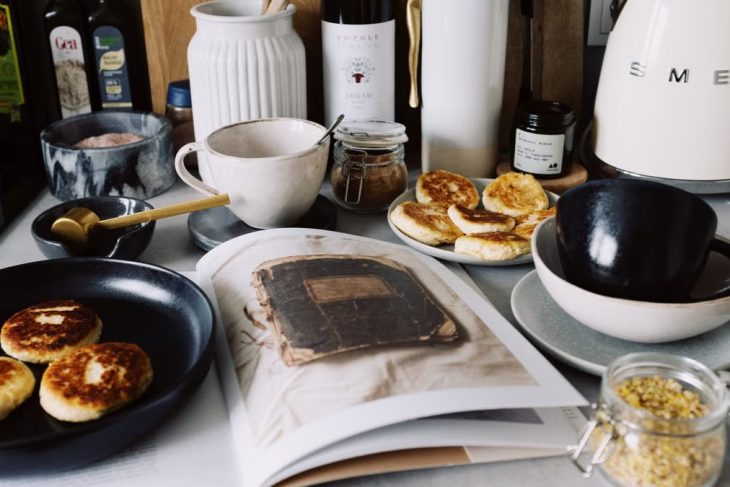 One portion of homemade cheese pancakes in frying pan and another on plate locating near opened stylish magazine among kitchen utensils and appliances in bright kitchen at home